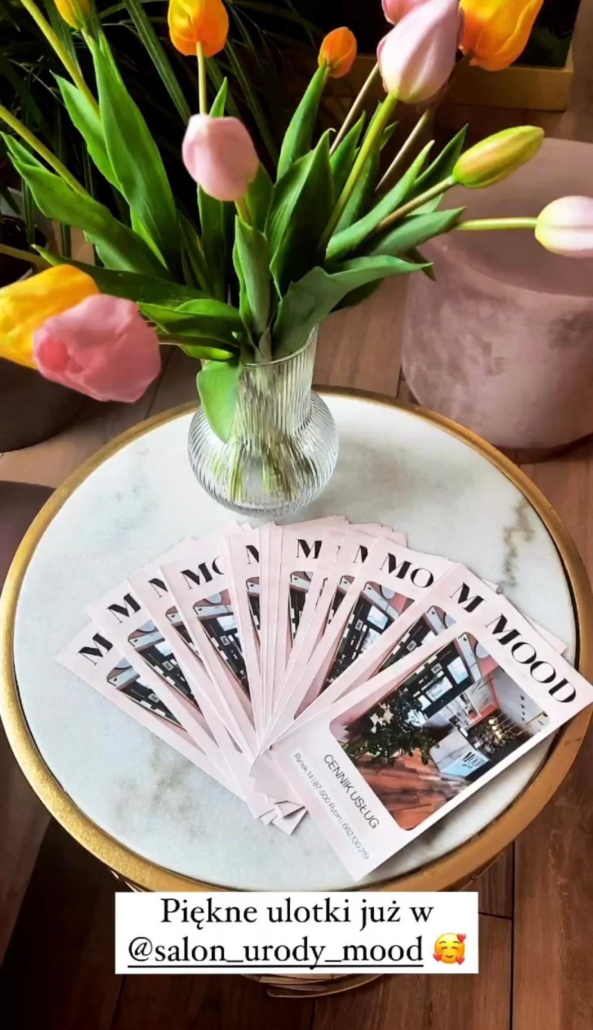 Stos różowych ulotek z cennikiem usług salonu urody Mood, ułożonych wachlarzowo na okrągłym stoliku z marmurowym blatem i złotą obwódką, obok wazon z tulipanami w odcieniach różu i żółci.