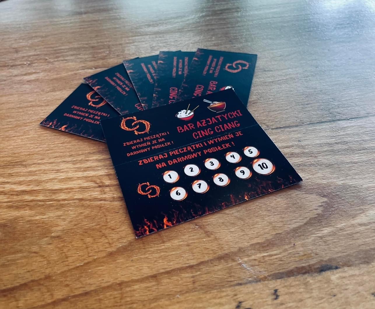 Stos czarnych kart lojalnościowych z logo i napisem 'Bar Azjatycki Cing Ciang' oraz informacją o zbieraniu pieczątek na darmowy posiłek, leżących na drewnianym stole.