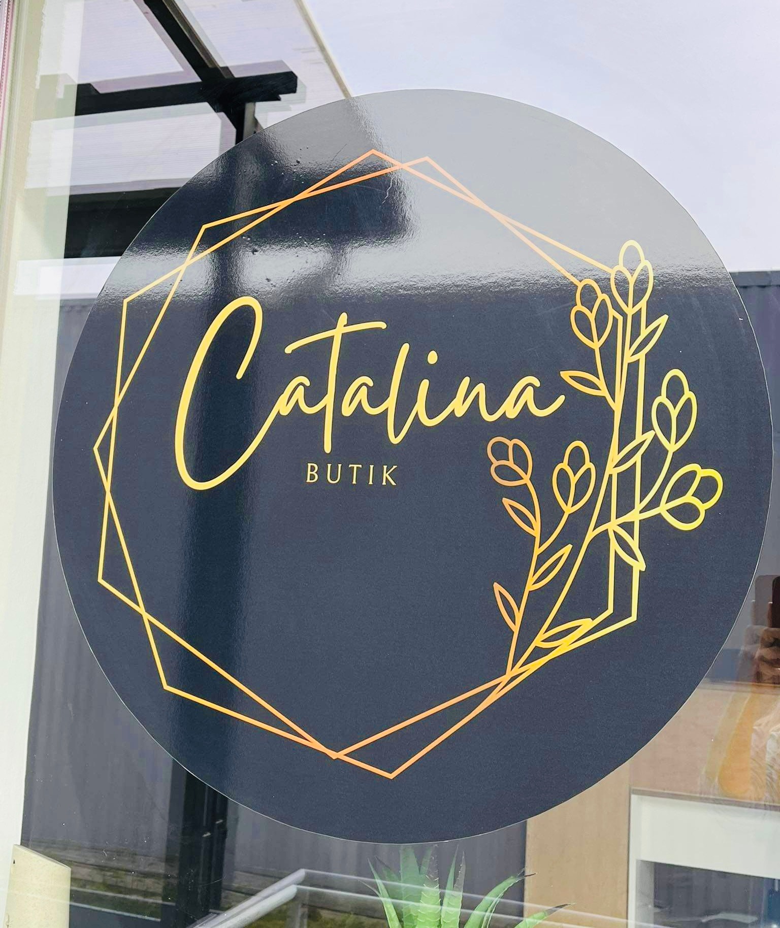 Okrągła naklejka z logo 'Catalina Butik' w kolorze złotym, umieszczona na szklanej witrynie, z elementami florystycznymi i geometrycznymi.