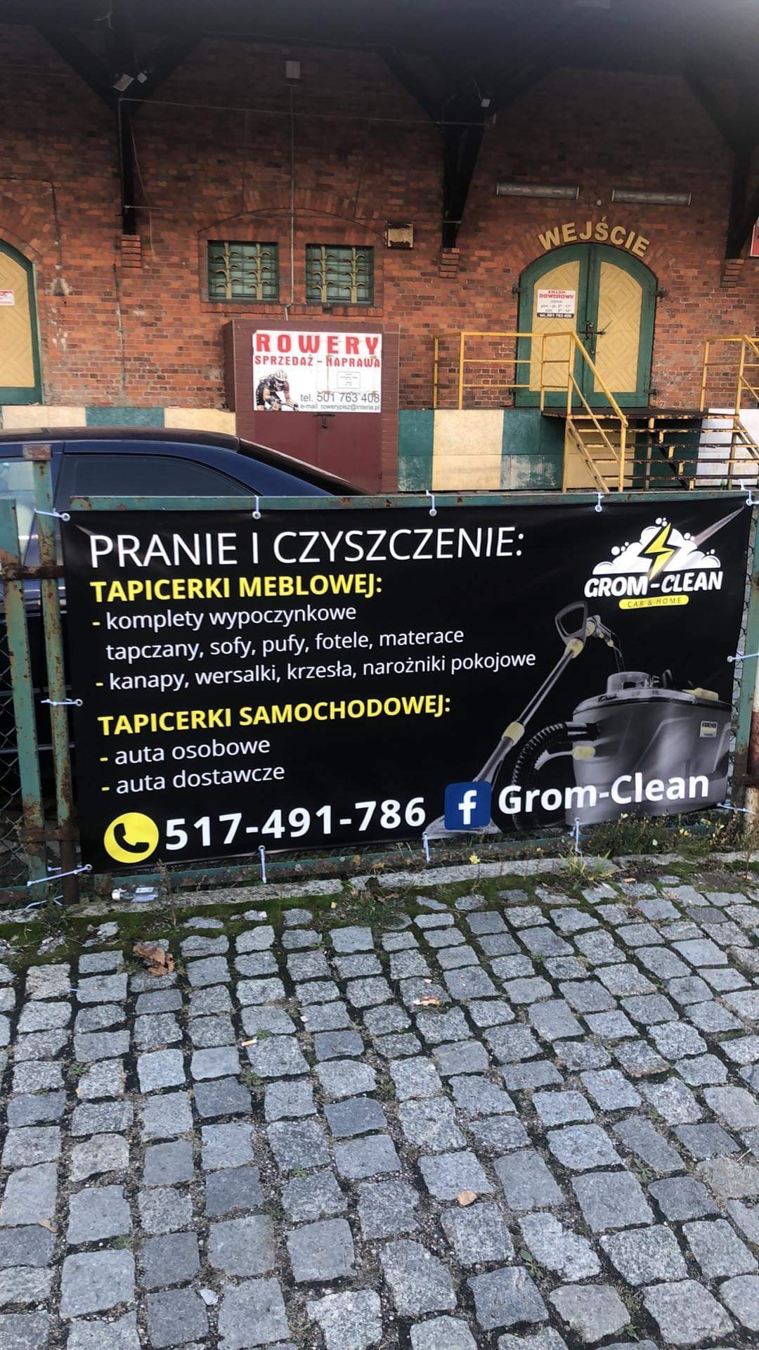 Baner reklamowy firmy Grom-Clean oferującej pranie i czyszczenie tapicerki meblowej i samochodowej, zawieszony na zewnątrz budynku z cegły.