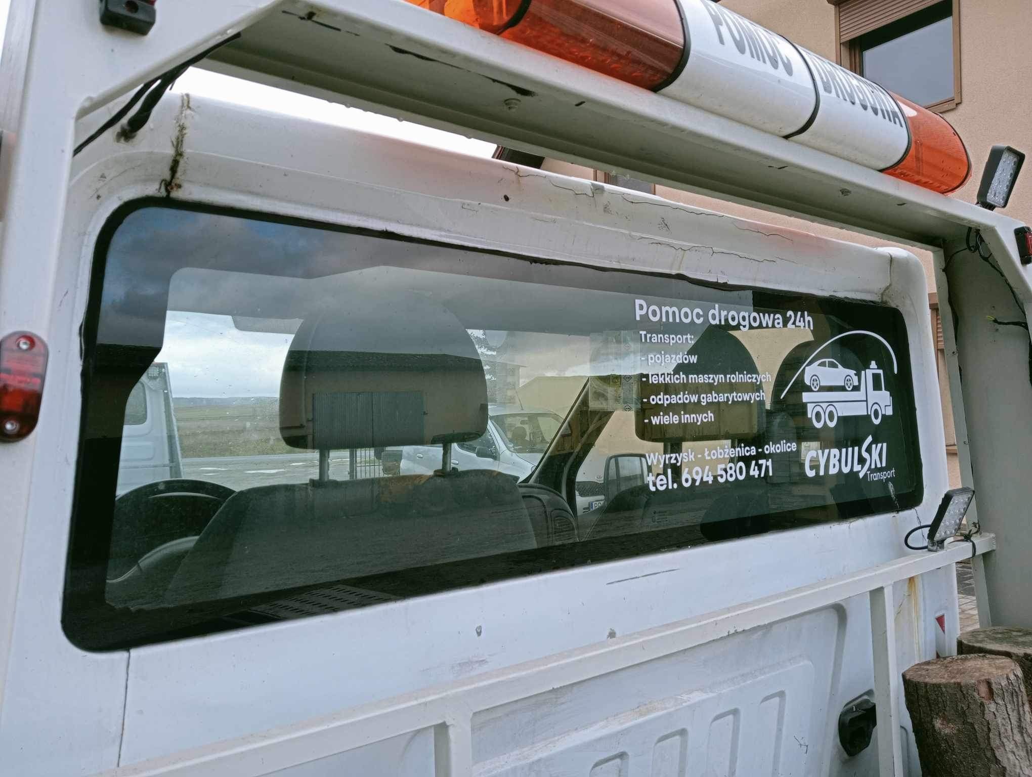Tylna szyba samochodu pomocy drogowej z naklejonymi informacjami kontaktowymi i zakresem usług, widoczne wnętrze kabiny i fragment sygnalizatora świetlnego na dachu.