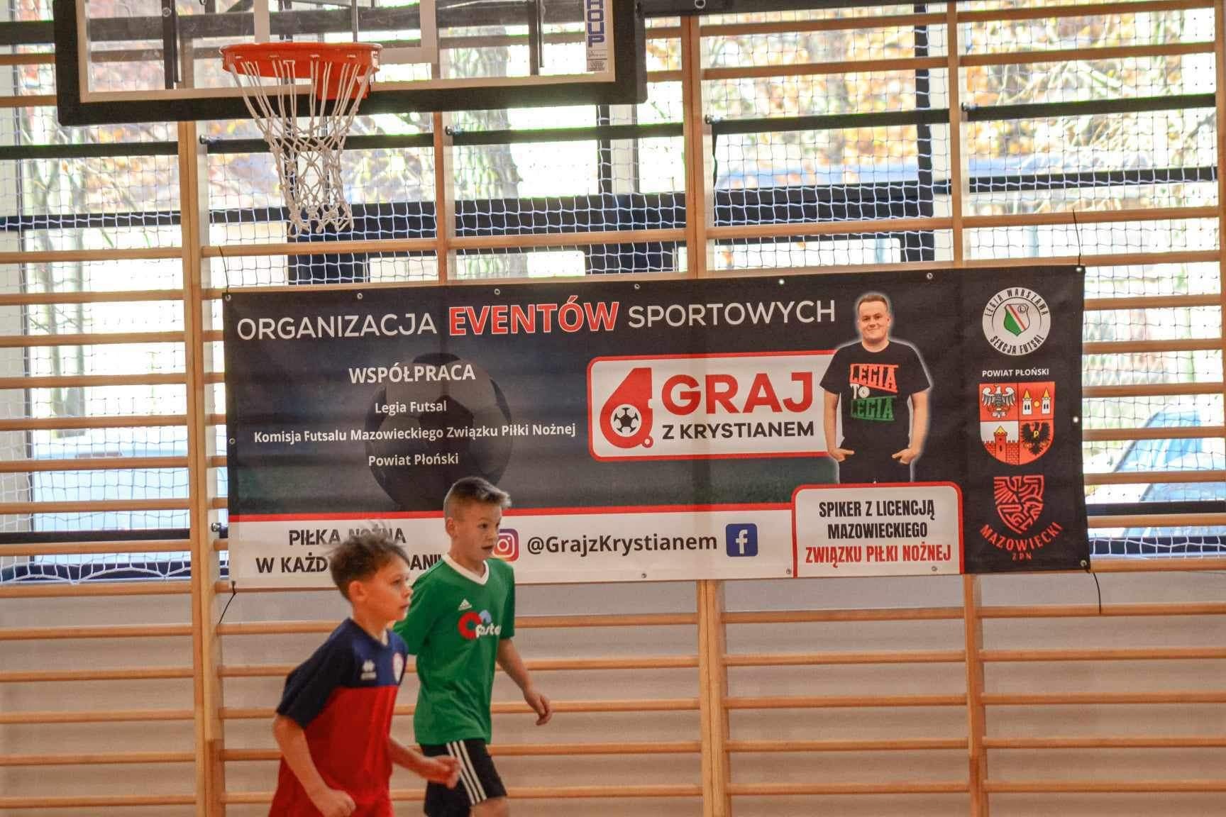 Sala gimnastyczna z widocznym koszem do koszykówki i banerem reklamującym organizację imprez sportowych, z dwójką dzieci w strojach sportowych w ruchu.