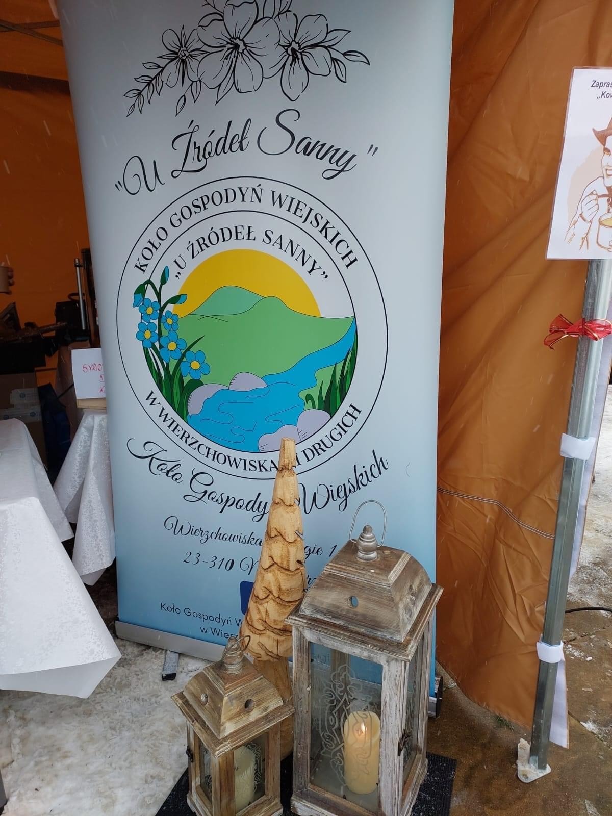 Stoisko Koła Gospodyń Wiejskich 'U Źródeł Sanny' z banerem reklamowym, drewnianą choinką i lampionami ze świecami.