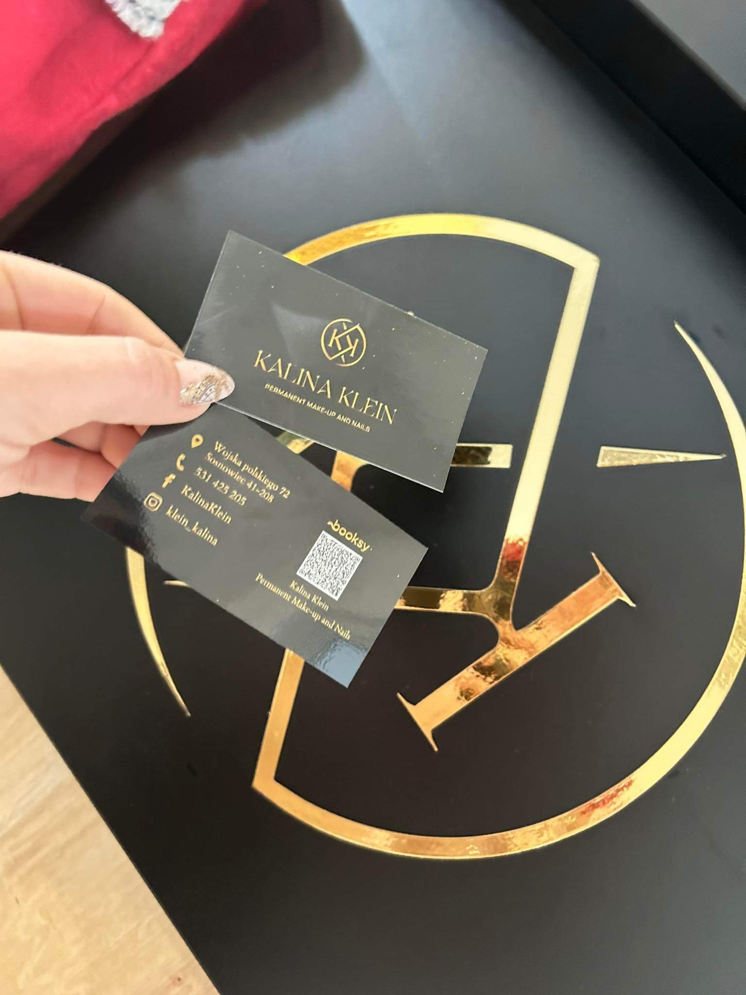 Dwie czarne wizytówki z błyszczącym złotym tekstem i kodem QR trzymane w dłoni na tle czarnej powierzchni z dużym złotym logo.