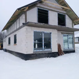Surowy stan domu w zimowej scenerii z częściowo zamontowanymi oknami PCV i przygotowaną konstrukcją dachu.