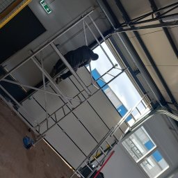 Sam21 - Montażysta na rusztowaniu instaluje białą bramę garażową segmentową w hali przemysłowej, widoczne narzędzia i części mechanizmu bramy.