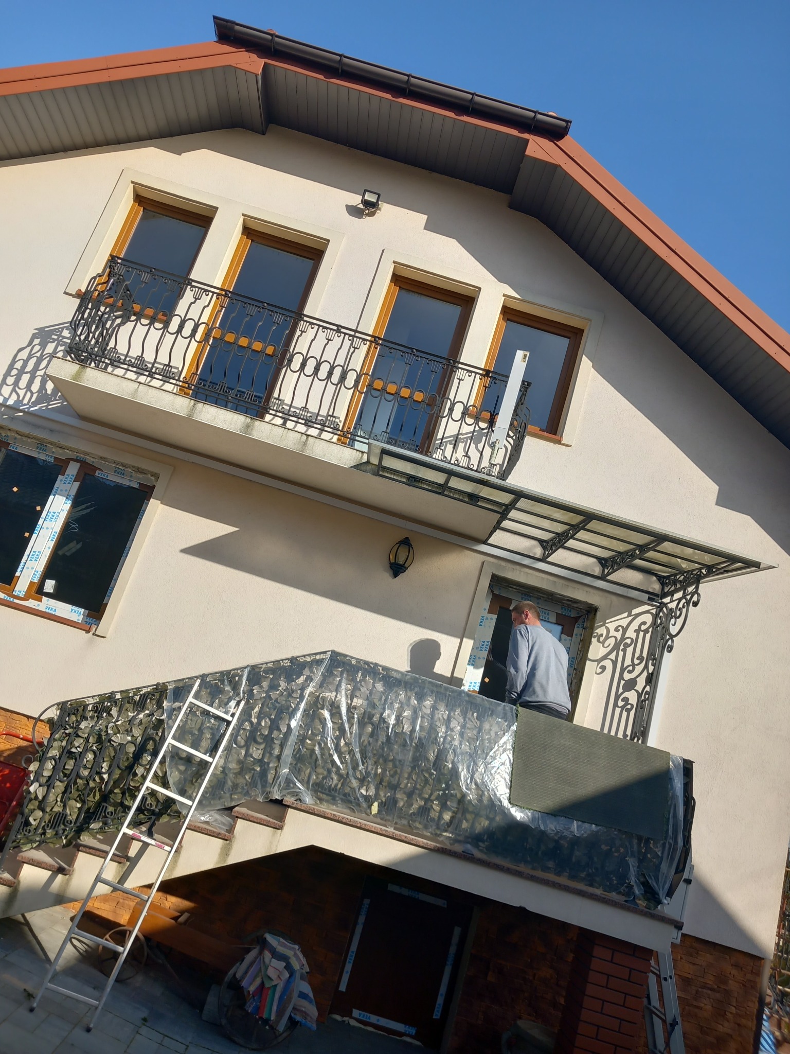 Montaż drzwi wejściowych pod ozdobnym zadaszeniem, widoczna drabina i folia zabezpieczająca, elewacja budynku w jasnym odcieniu.