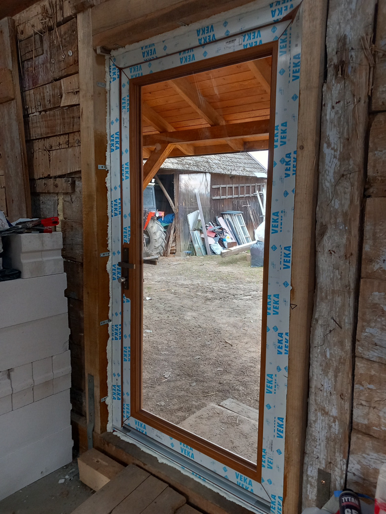 Montaż brązowych drzwi zewnętrznych z folią ochronną VEKA na ościeżnicy, widok z wnętrza budynku gospodarczego z drewnianymi ścianami, w tle traktor i sterta starych okien.