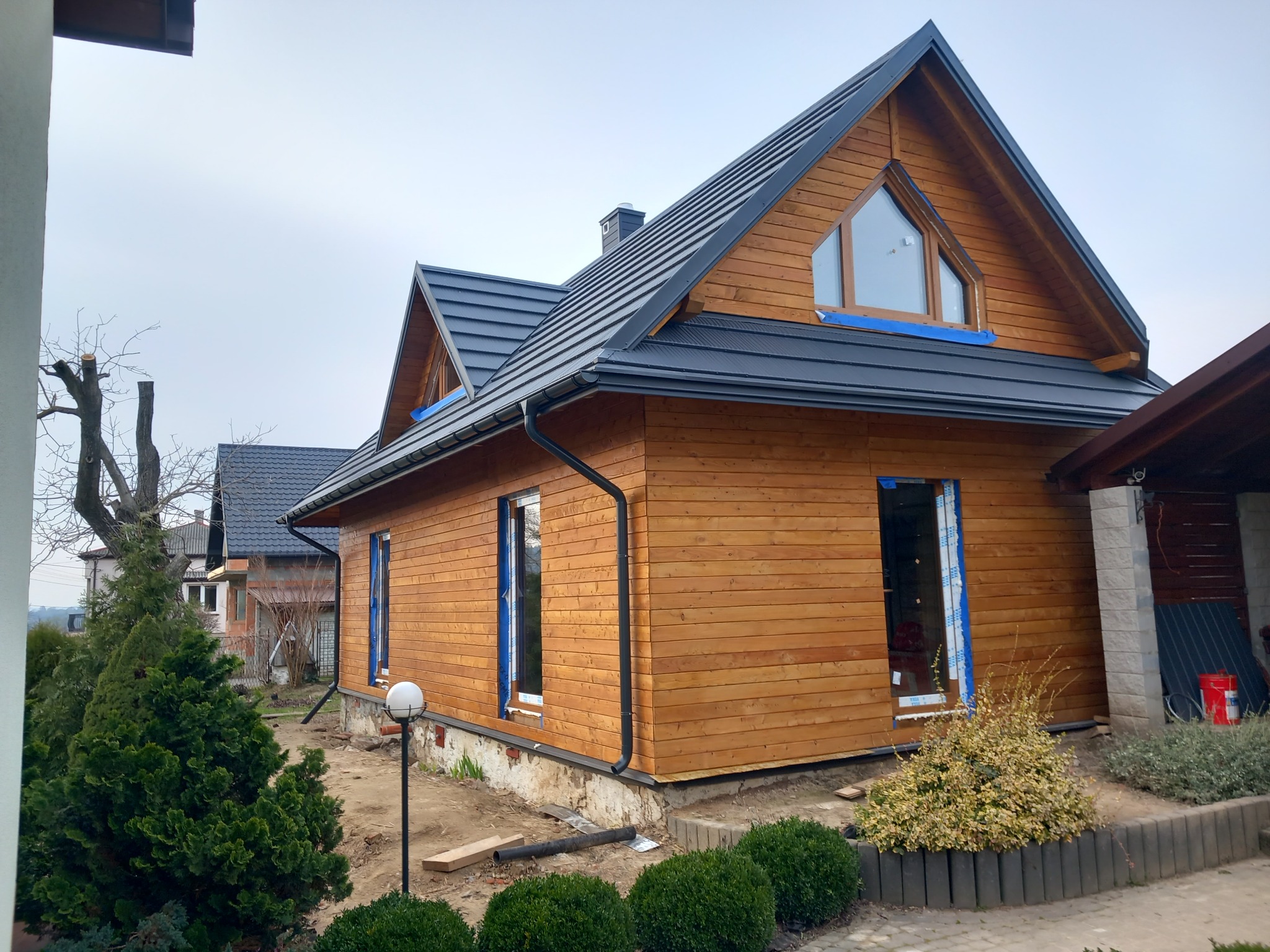 Drewniany dom w trakcie budowy lub wykończenia, widoczne zabezpieczenia okien folią, czarna dachówka i drewniana elewacja, oświetlenie zewnętrzne w formie kuli.