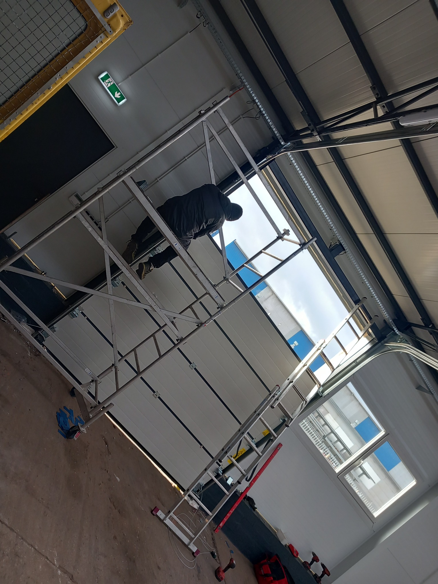 Montażysta na rusztowaniu instaluje białą bramę garażową segmentową w hali przemysłowej, widoczne narzędzia i części mechanizmu bramy.