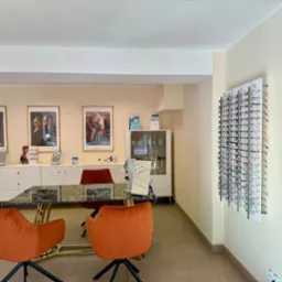 Salon optyczny: pomarańczowe krzesła przy stole z marmurowym blatem, ekspozycja oprawek na ścianie, plakaty reklamowe na szafce. Jasne wnętrze.