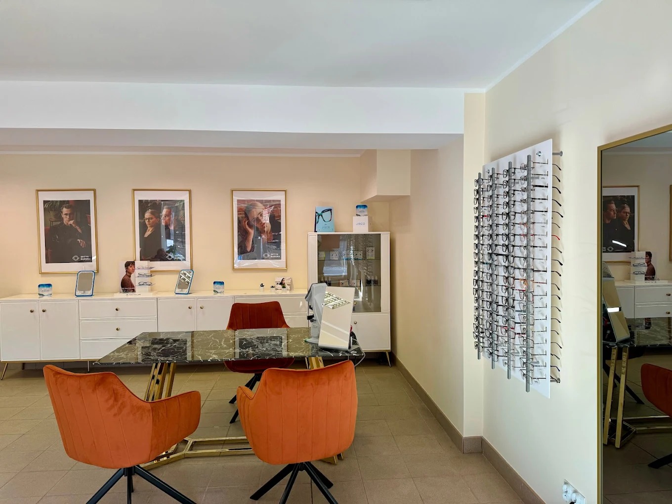 Salon optyczny: pomarańczowe krzesła przy stole z marmurowym blatem, ekspozycja oprawek na ścianie, plakaty reklamowe na szafce. Jasne wnętrze.