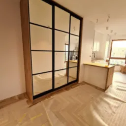 Duża szafa z lustrzanymi drzwiami w stylu loft, z czarnymi szprosami, w trakcie montażu; podłoga zabezpieczona folią malarską, widoczna kuchnia w tle.