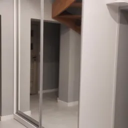 Biała szafa wnękowa z przesuwnymi, lustrzanymi drzwiami w mieszkaniu w Wołowie, odbijająca drewniane schody i drzwi w tle.