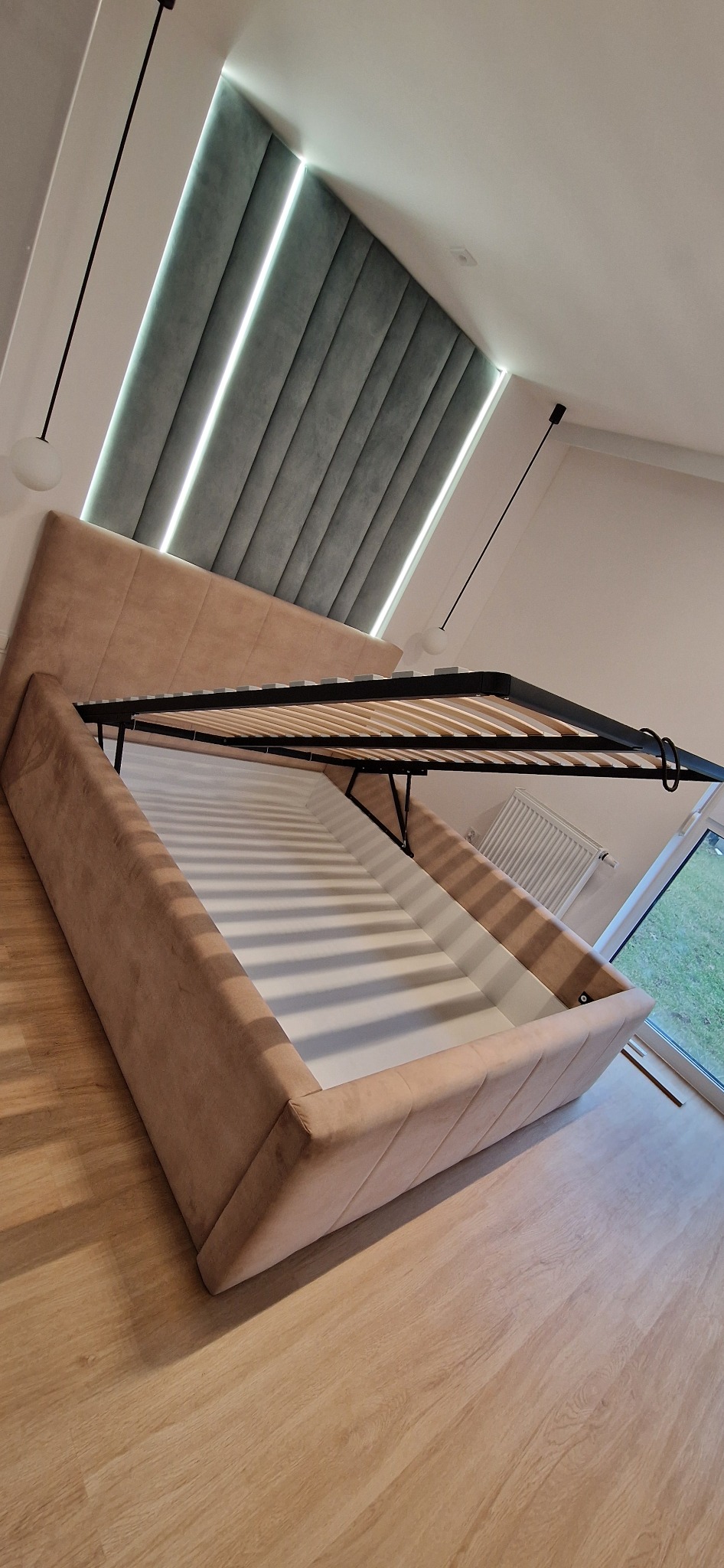 Tapicerowane łóżko z podnoszonym stelażem i pojemnikiem na pościel, widok z dołu, na podłodze panele, w tle zagłówek z oświetleniem.