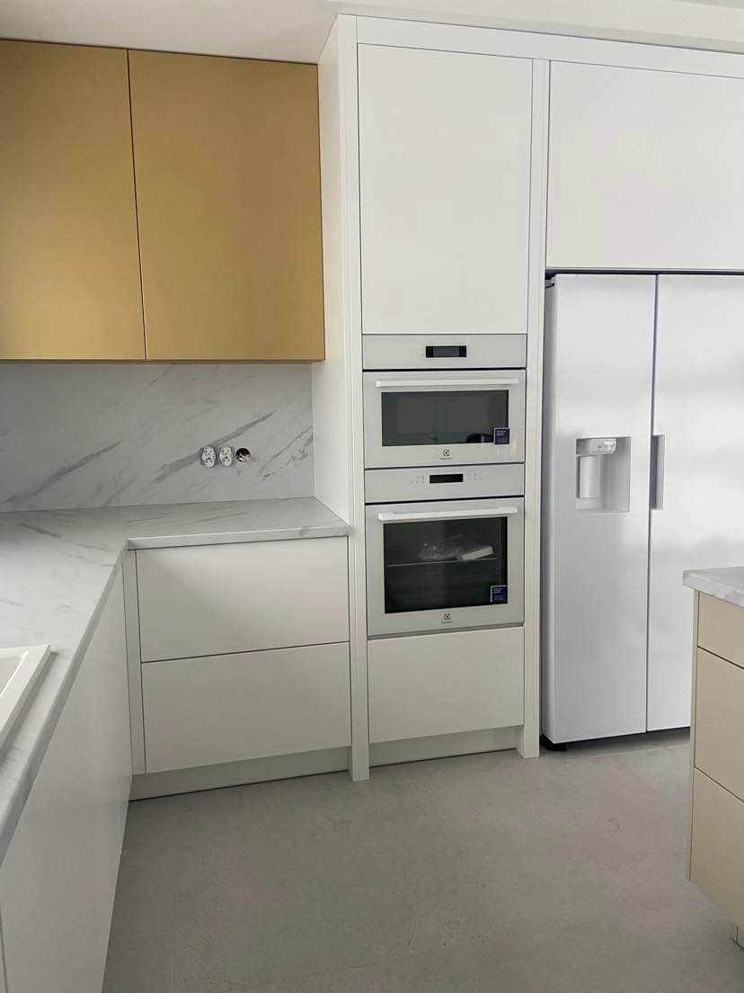 Nowoczesna kuchnia z białymi szafkami, złotymi górnymi szafkami, marmurowym blatem i wbudowanym sprzętem AGD marki Electrolux, w tym piekarnik i lodówka side-by-side.