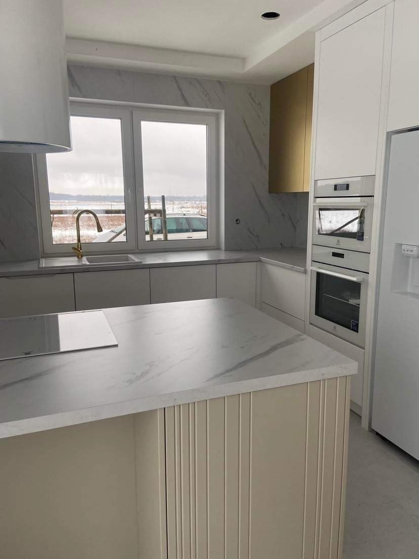 Nowoczesna kuchnia z wyspą, białymi szafkami i blatem imitującym marmur, złota bateria przy oknie z widokiem na zimowy krajobraz.