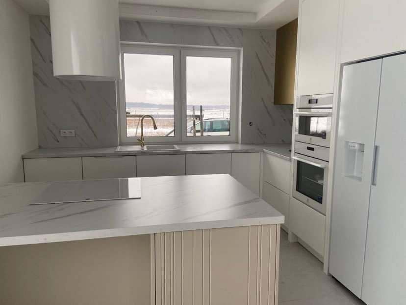 Nowoczesna, jasna kuchnia z wyspą, białymi frontami i blatem imitującym marmur, złota bateria przy oknie z widokiem na krajobraz za oknem.