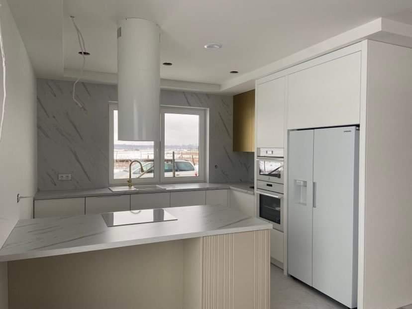 Nowoczesna, minimalistyczna kuchnia w odcieniach bieli i szarości z wyspą kuchenną, marmurkowymi blatami i frontami szafek, oraz złotym akcentem w górnej szafce, widoczna przez okno zimowa sceneria.