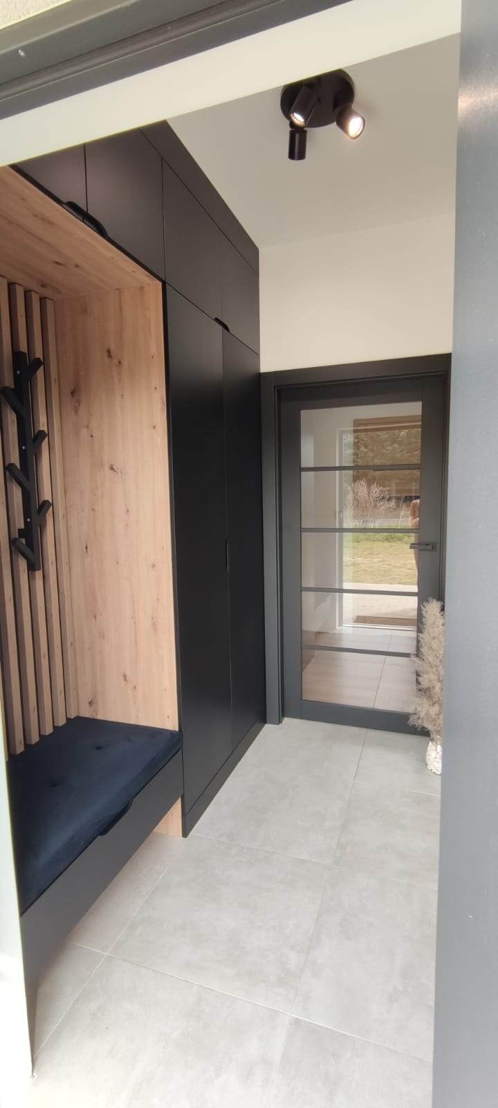 Nowoczesny przedpokój z zabudową: czarne szafy, drewniany panel z wieszakami i siedziskiem z granatową poduszką, szklane drzwi w czarnej ramie.