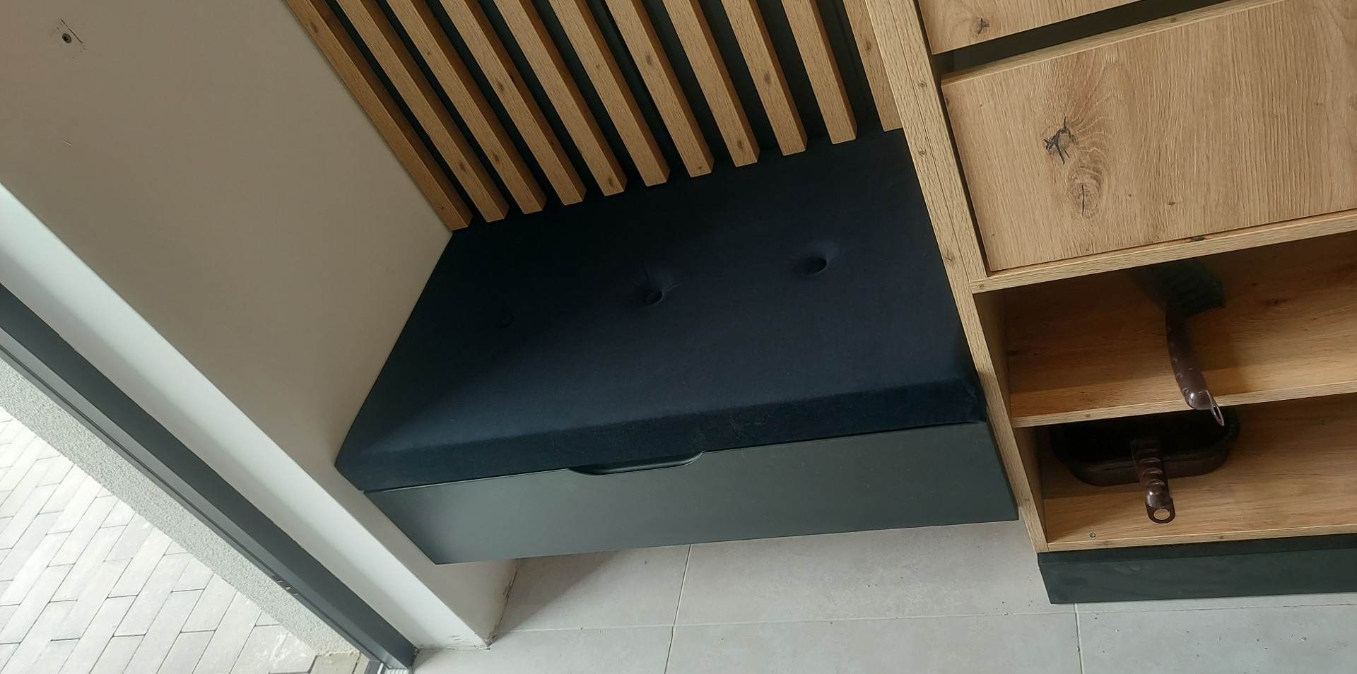 Wnęka z siedziskiem z ciemną tapicerką i drewnianymi lamelami na ścianie, obok szafka z drewna z widocznymi naczyniami w środku.