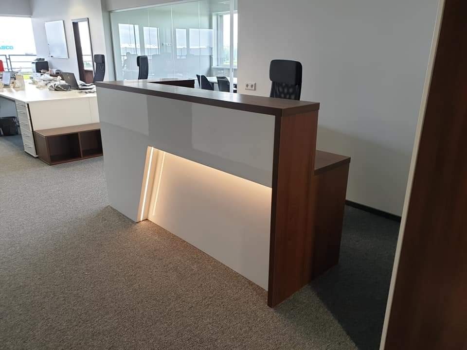 Nowoczesna lada recepcyjna z białymi panelami i drewnianymi elementami, podświetlana od spodu ciepłym światłem LED, na tle biura z przeszkloną ścianą i stanowiskami pracy.