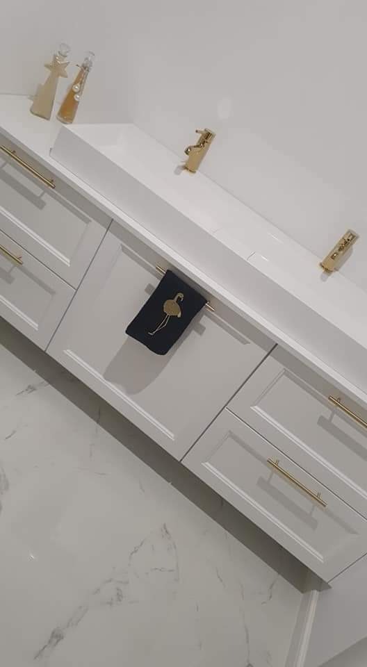 Szare szafki z frezowanymi frontami i złotymi uchwytami, czarna ściereczka z motywem flaminga zawieszona na uchwycie, biały blat z dekoracyjnymi butelkami i złotą armaturą, marmurowa podłoga.