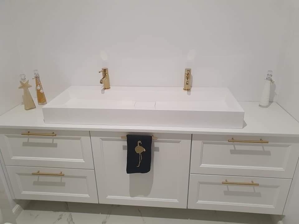 Biała, prostokątna umywalka z dwoma złotymi bateriami, osadzona na szafce z białymi frontami i złotymi uchwytami. Na środkowych drzwiczkach szafki wisi czarny ręcznik z haftowanym złotym flamingiem...