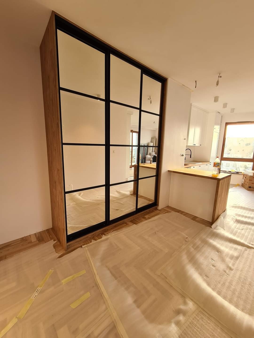 Duża szafa z lustrzanymi drzwiami w stylu loft, z czarnymi szprosami, w trakcie montażu; podłoga zabezpieczona folią malarską, widoczna kuchnia w tle.