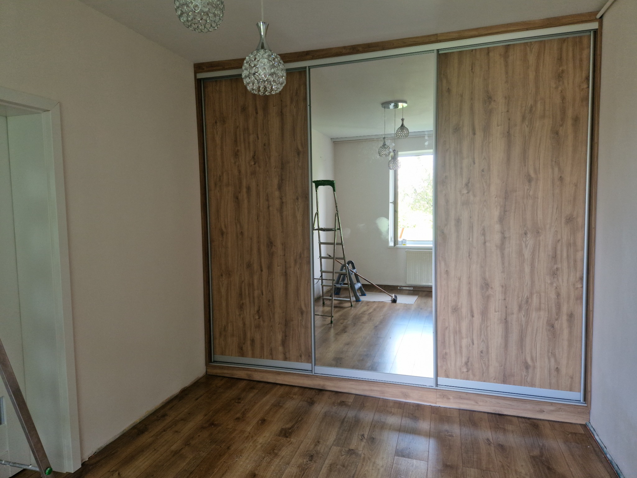 Wnętrze pokoju z dużą szafą przesuwną z lustrzanymi drzwiami i drewnopodobnymi panelami, widoczna drabina malarska w odbiciu lustra, drewniana podłoga.