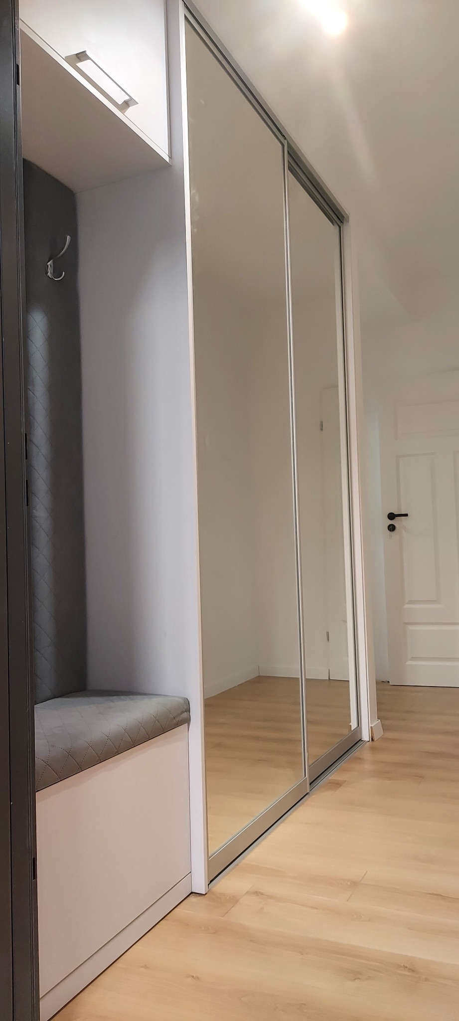 Wnęka z siedziskiem z szarej pikowanej tkaniny i haczykiem na ścianie, obok szafa wnękowa z lustrzanymi przesuwnymi drzwiami, widoczna podłoga z paneli.