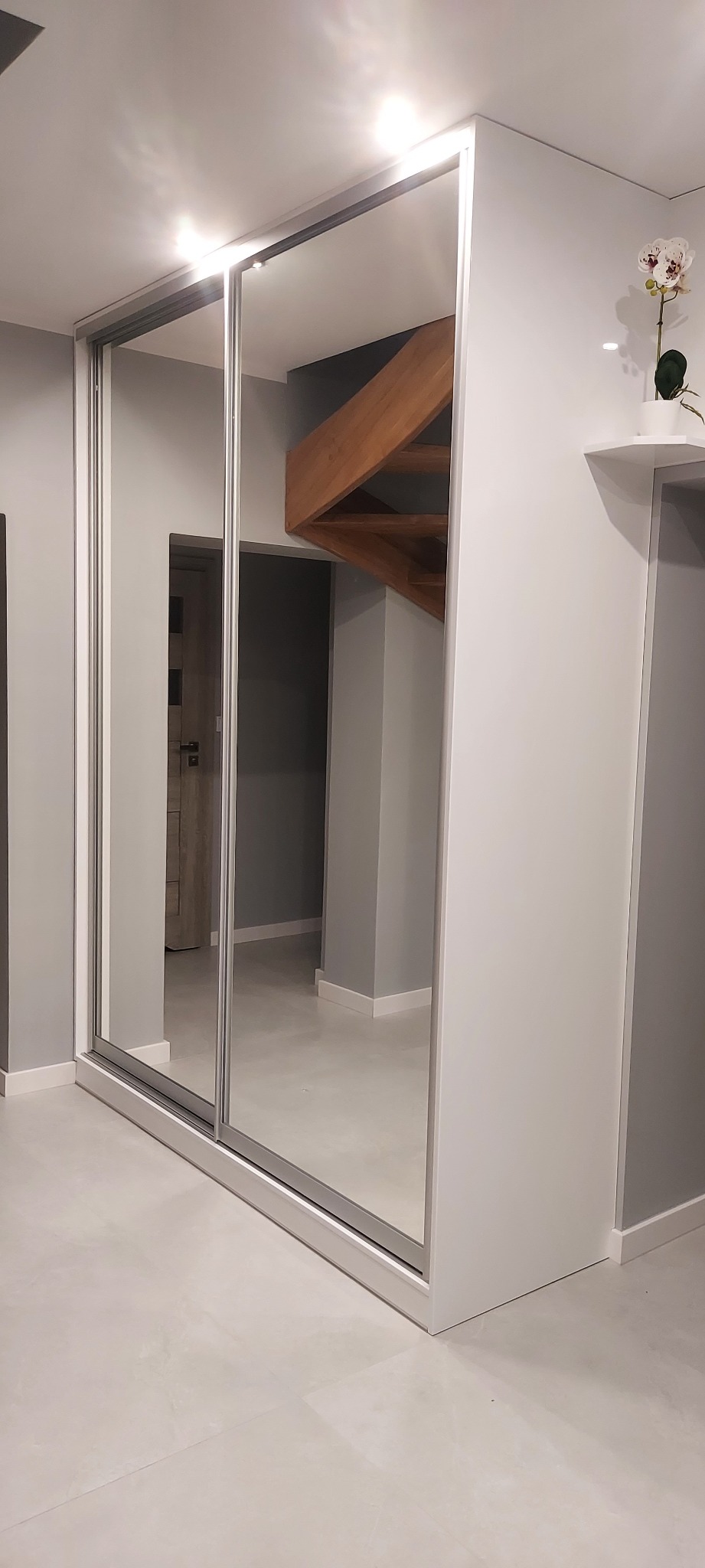 Biała szafa wnękowa z przesuwnymi, lustrzanymi drzwiami w mieszkaniu w Wołowie, odbijająca drewniane schody i drzwi w tle.