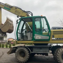 Żółto-zielona koparka kołowa Liebherr 900 Litronic z podniesioną łyżką, widoczna na tle domu i drzew bez liści w pochmurny dzień.