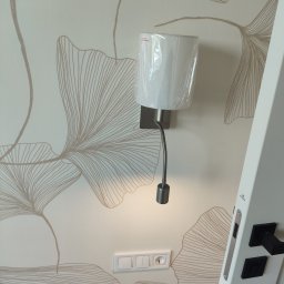 Viktor Drozdov Elektryk - Świecąca lampka nocna LED z elastycznym ramieniem, zamontowana na ścianie z motywem liści, obok włącznika i fragmentu białych drzwi z czarną klamką.