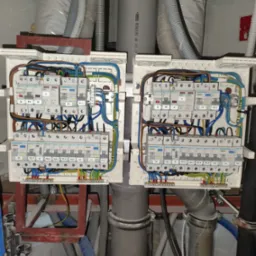 Dwa białe panele elektryczne z zainstalowanymi bezpiecznikami i okablowaniem, widoczne rury i przewody instalacyjne w tle.