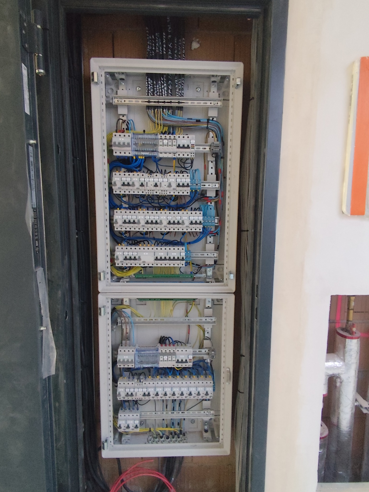 Dwu-segmentowa, otwarta skrzynka elektryczna z licznymi bezpiecznikami i okablowaniem, zamontowana w ścianie, widoczne przewody i elementy instalacji.