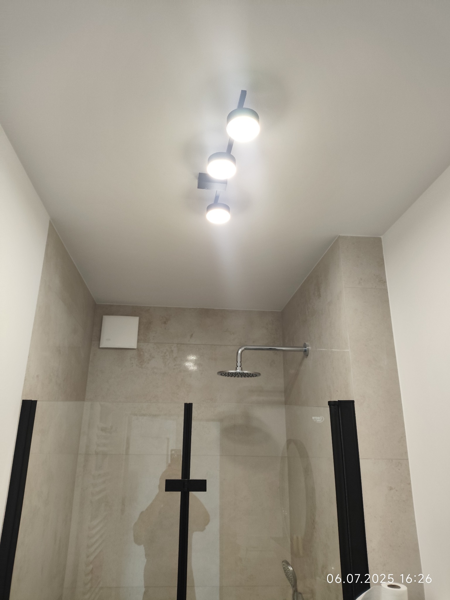 Nowoczesna kabina prysznicowa z czarnymi profilami i zamontowanym oświetleniem punktowym na suficie, wykończona beżowymi płytkami. Widoczny deszczownica.