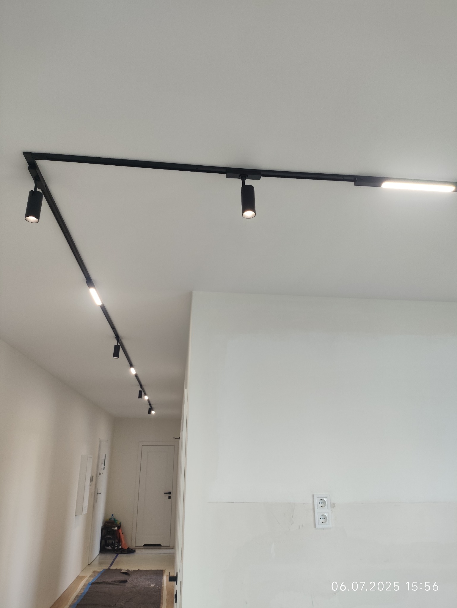 Czarne szynoprzewody z reflektorami i listwą LED, zamontowane na białym suficie w korytarzu. Widoczne gniazdka elektryczne na ścianie.