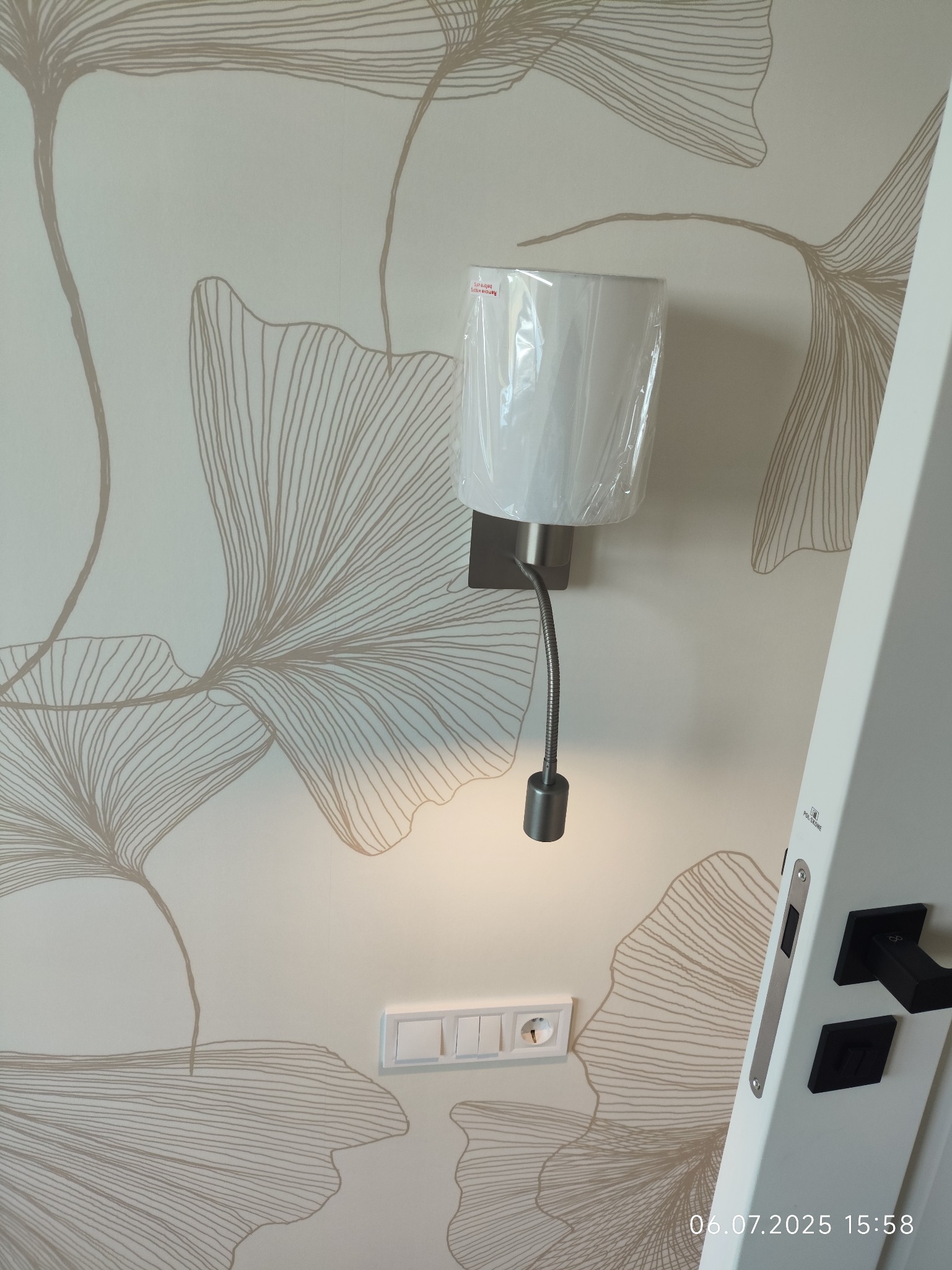 Świecąca lampka nocna LED z elastycznym ramieniem, zamontowana na ścianie z motywem liści, obok włącznika i fragmentu białych drzwi z czarną klamką.