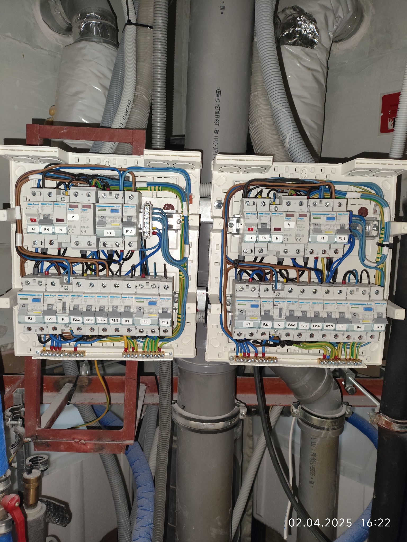 Dwa białe panele elektryczne z zainstalowanymi bezpiecznikami i okablowaniem, widoczne rury i przewody instalacyjne w tle.