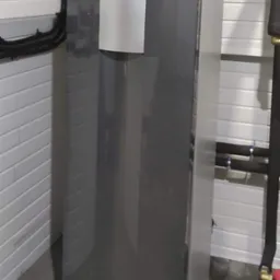 Szara pompa ciepła zainstalowana w pomieszczeniu z białą boazerią, widoczne przyłącza rur w izolacji oraz panel sterowania z zielonym przyciskiem.