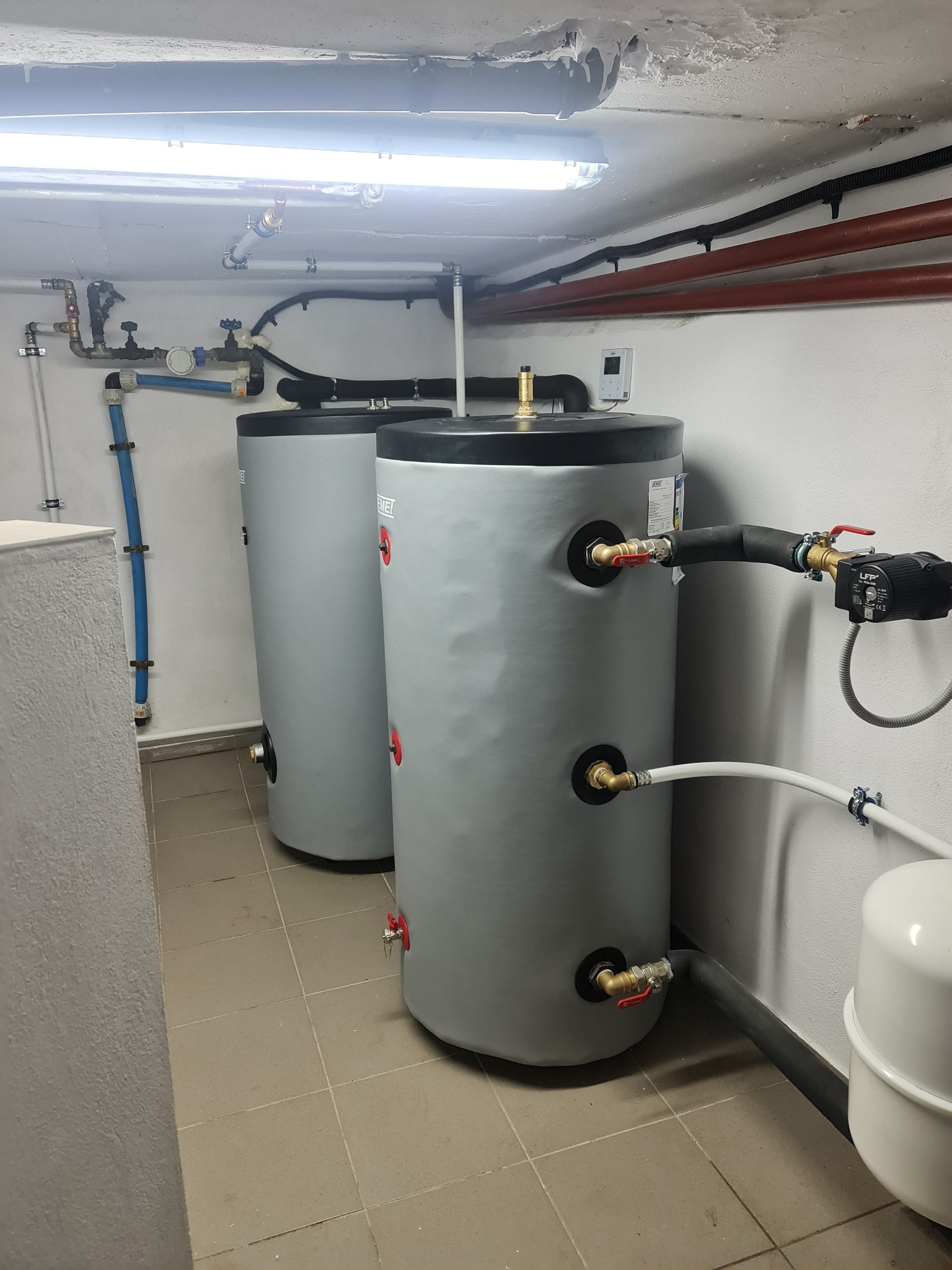 Dwa szare, cylindryczne zbiorniki na wodę w kotłowni, z widocznymi rurami, zaworami i pompą obiegową, na tle białych ścian i ceramicznej podłogi.