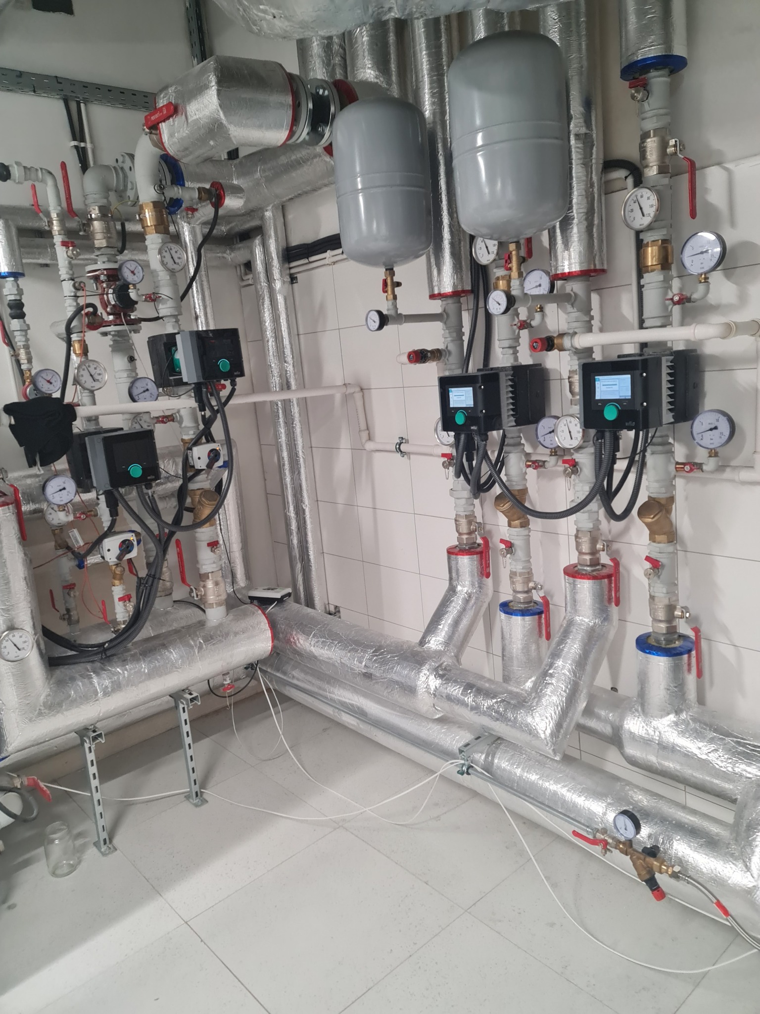Węzeł cieplny z izolowanymi rurami, manometrami, zaworami i sterownikami elektronicznymi w pomieszczeniu z białymi płytkami.
