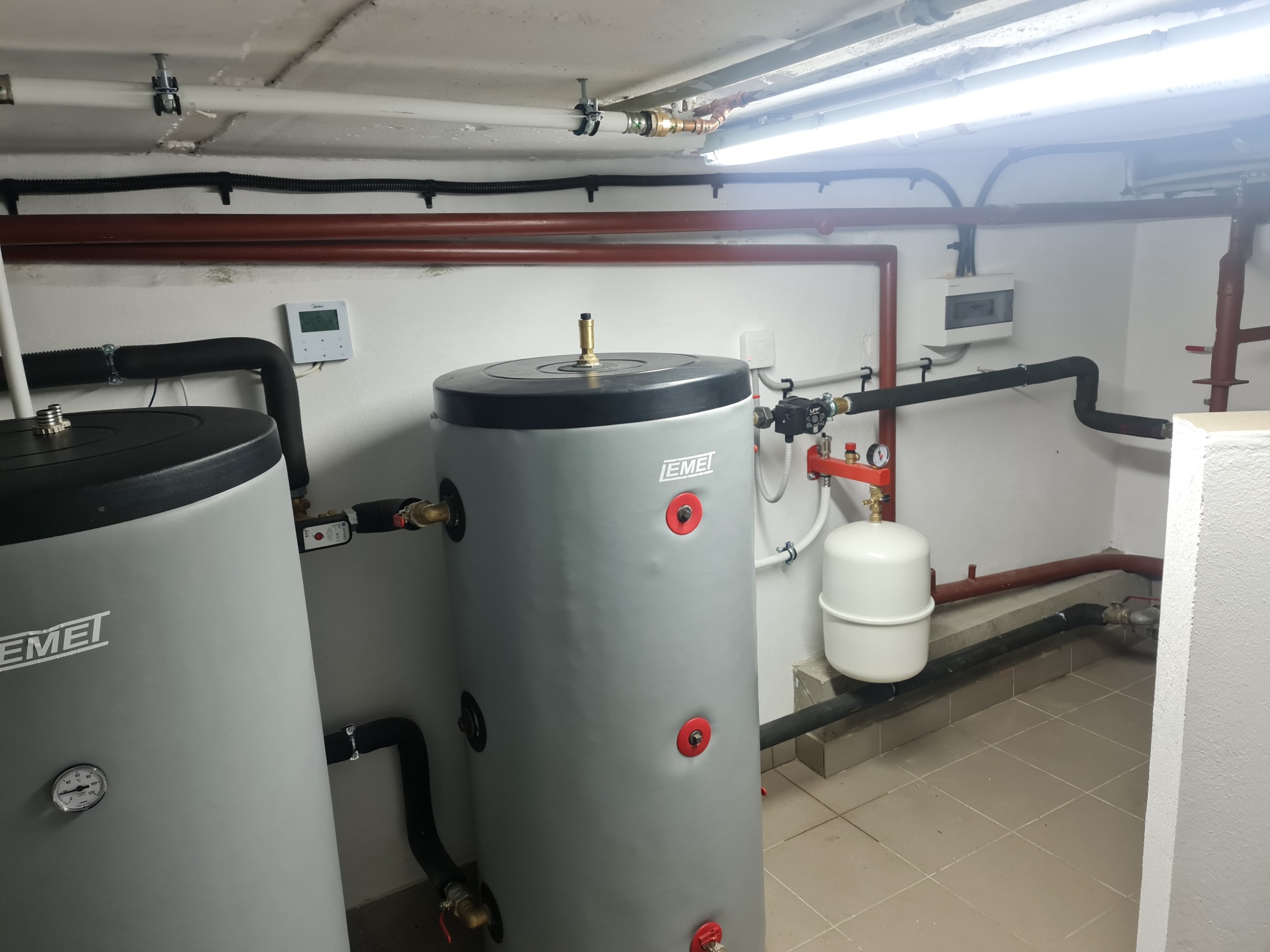 Dwa szare zasobniki ciepłej wody użytkowej marki EMEI w kotłowni, widoczne rury, manometr, biały zbiornik wyrównawczy, płytki na podłodze i sterownik na ścianie.