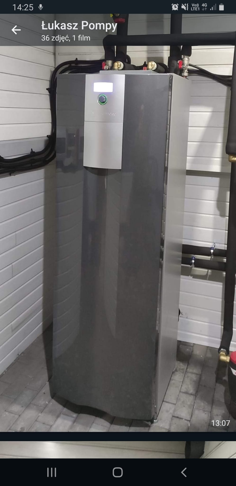 Szara pompa ciepła zainstalowana w pomieszczeniu z białą boazerią, widoczne przyłącza rur w izolacji oraz panel sterowania z zielonym przyciskiem.