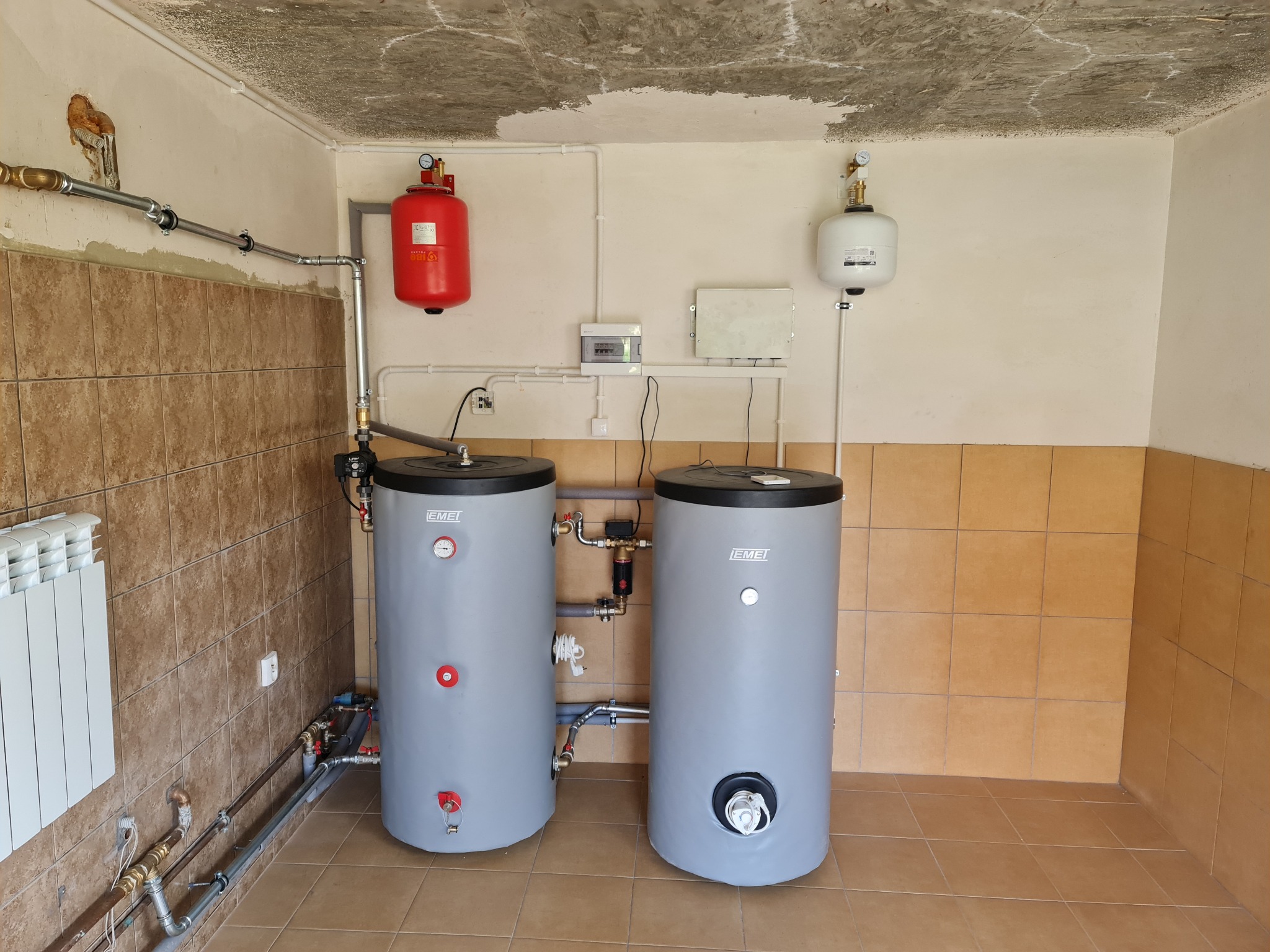 Dwa szare zbiorniki akumulacyjne ciepła marki EMET w kotłowni z brązowymi płytkami, widoczne rury instalacji CO, czerwony i biały zbiornik wyrównawczy oraz surowy betonowy sufit.
