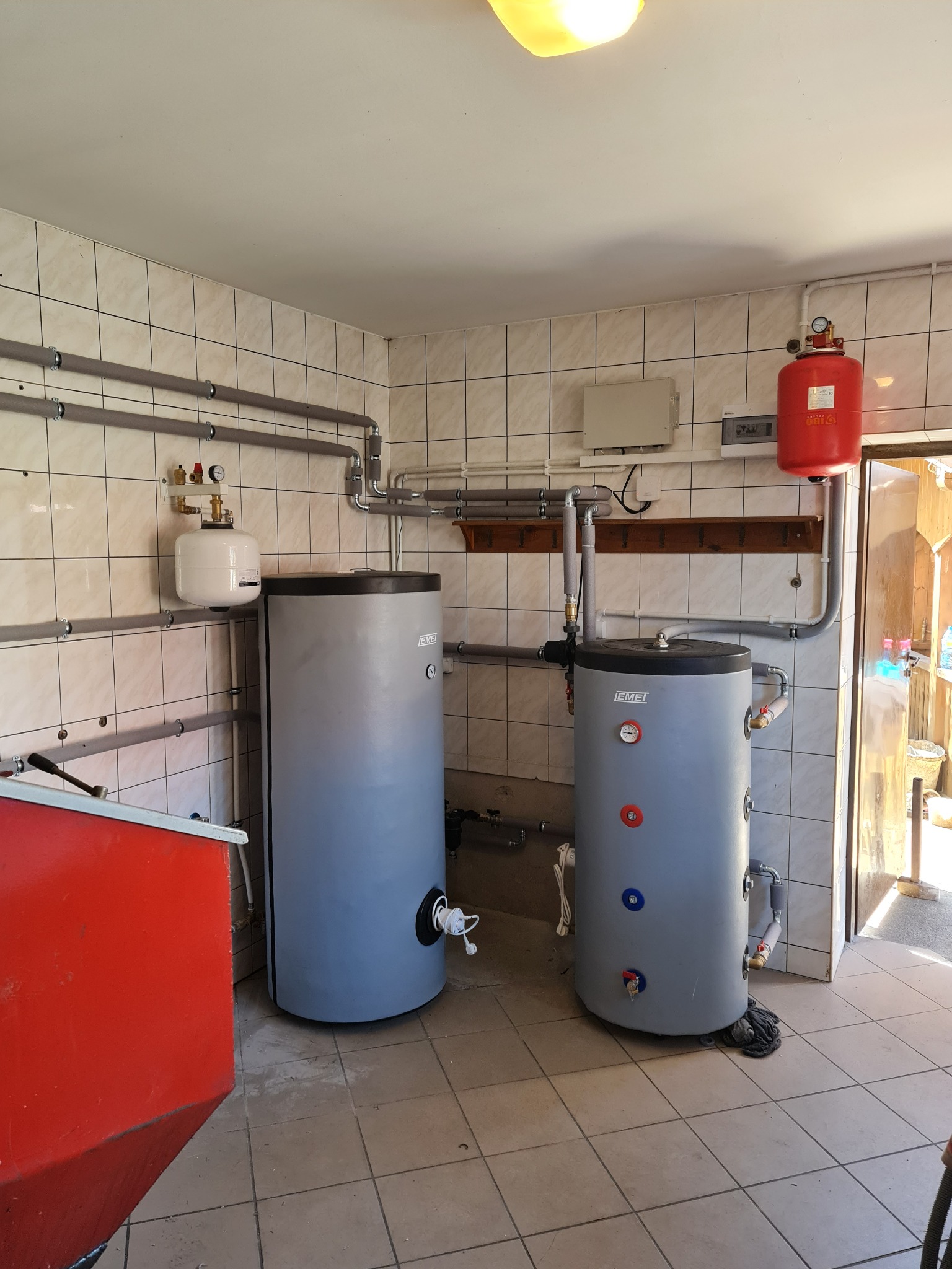 Dwa szaro-niebieskie zasobniki ciepłej wody użytkowej firmy Emei w kotłowni z ceramicznymi płytkami na ścianach, widoczne rury instalacyjne z izolacją, czerwony zbiornik wyrównawczy oraz fragment...