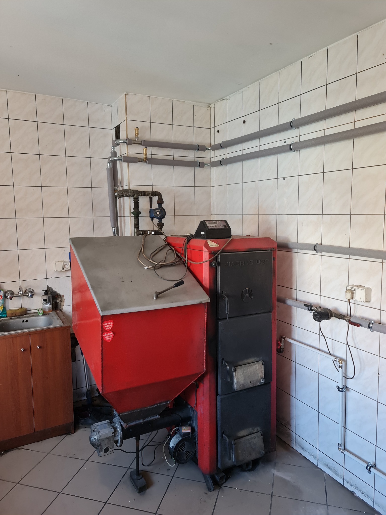 Czerwono-czarny piec na paliwo stałe Viadrus U22 z podajnikiem, zainstalowany w kotłowni wyłożonej białymi płytkami, widoczne rury centralnego ogrzewania z izolacją.