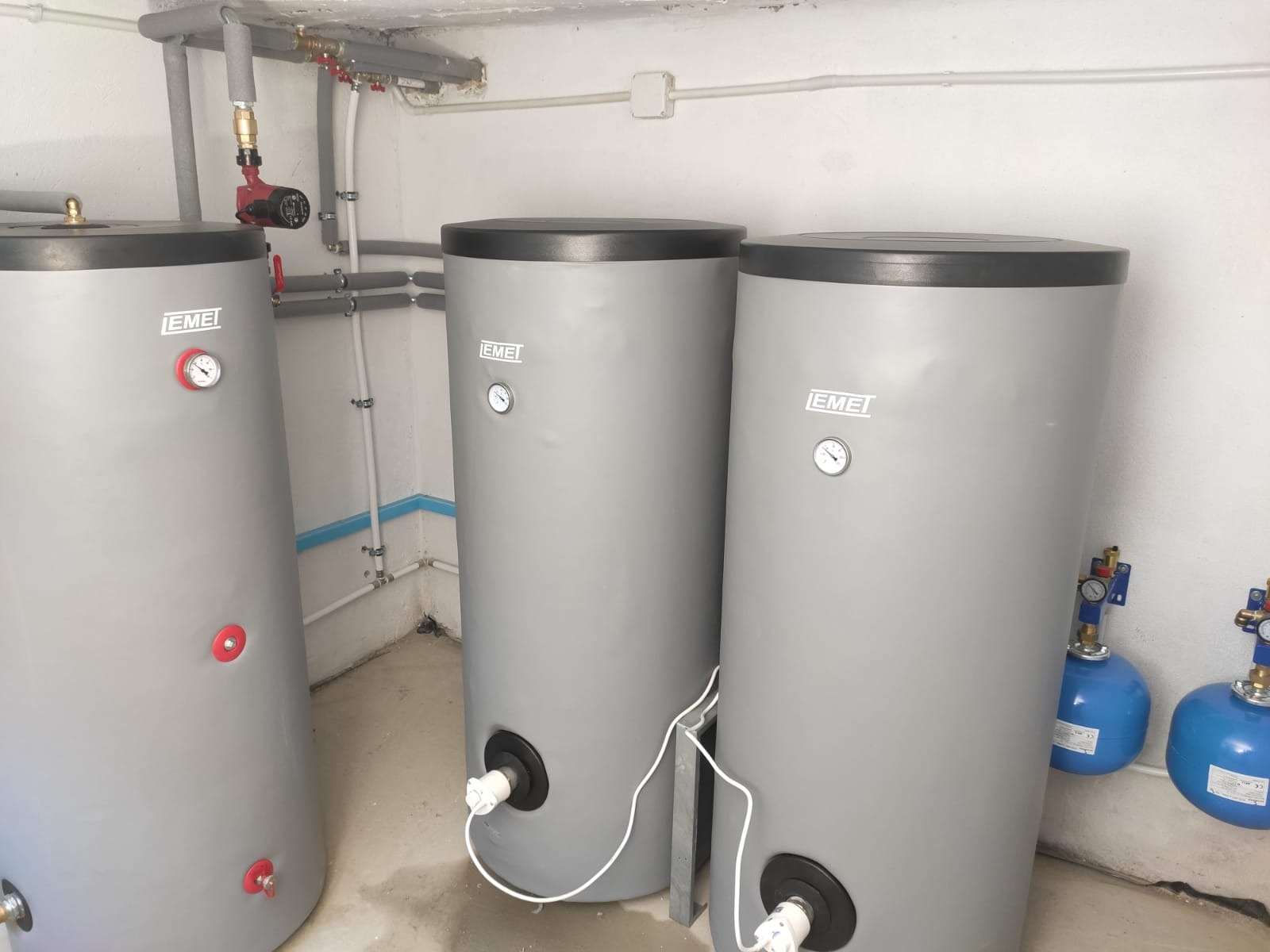 Trzy szare zasobniki ciepłej wody użytkowej marki EMEI z podłączonymi rurami i osprzętem w pomieszczeniu technicznym.