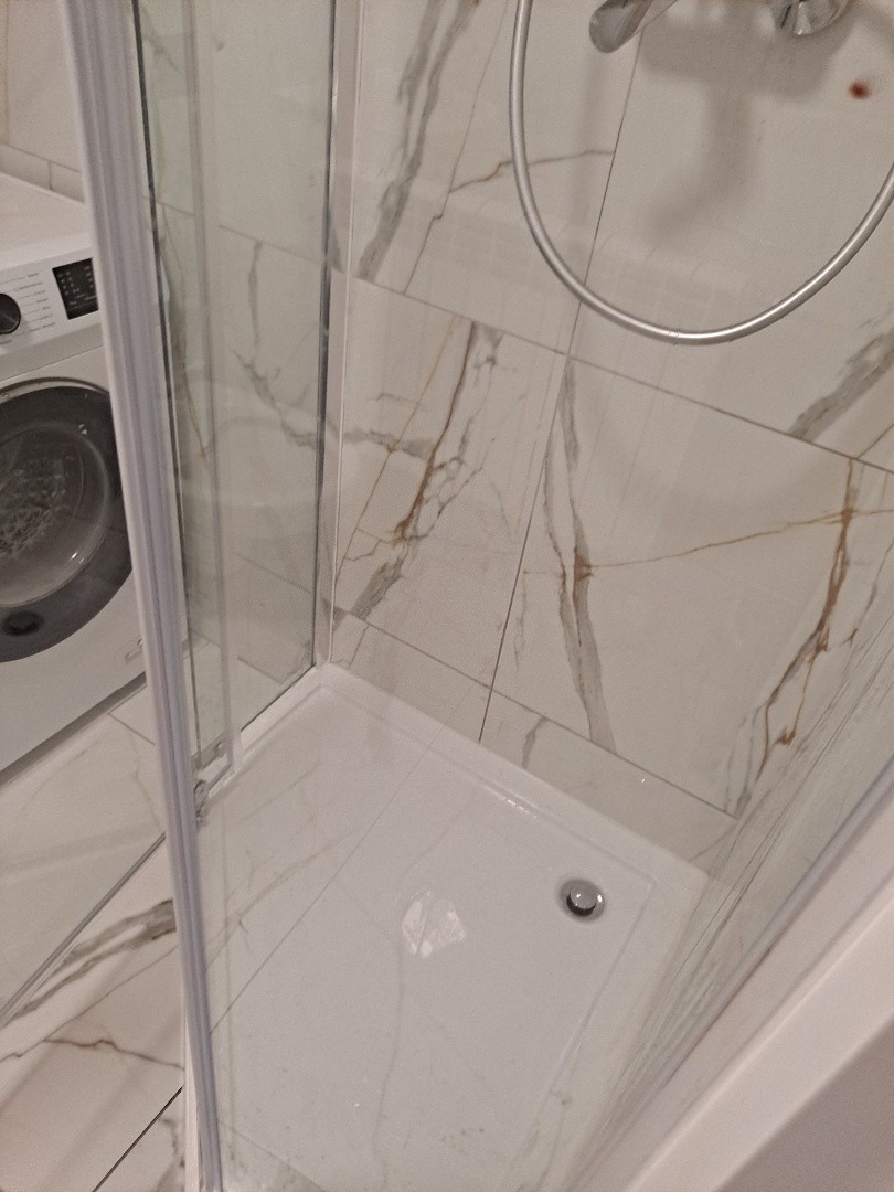 Biały brodzik kabiny prysznicowej z odpływem, otoczony marmurowymi płytkami z brązowymi żyłkami. W tle pralka i chromowana rura natrysku.